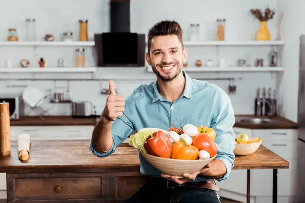 Joven feliz sosteniendo tazón con verduras frescas y mostrando el pulgar en la cocina - foto de stock