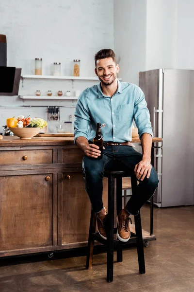 Handsome young man holding bottle of beer and smiling at camera in kitchen — Stock Photo