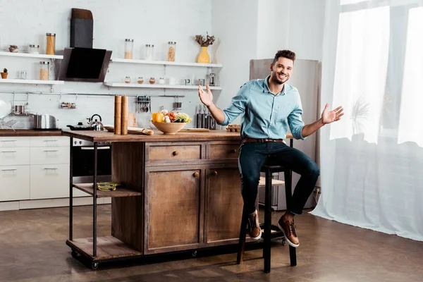 Fröhlicher junger Mann lächelt in die Kamera, während er mit offenen Armen in der Küche sitzt — Stockfoto