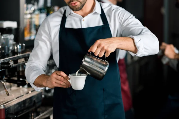 Vista recortada de barista en delantal vertiendo leche al café en el lugar de trabajo - foto de stock