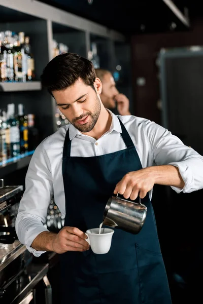 Bello barista in grembiule versando latte dal barattolo sul posto di lavoro — Foto stock