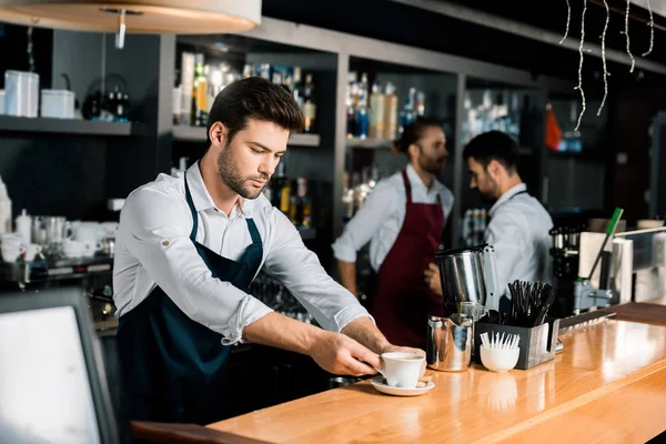 Bel barista in grembiule mettere tazza di caffè al bancone di legno — Foto stock