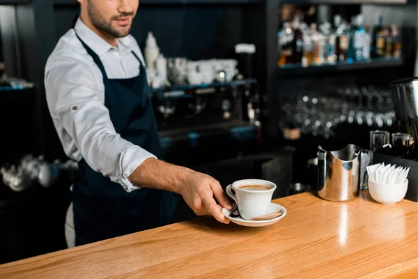 Vista recortada de barista en delantal poniendo taza de café en mostrador de madera - foto de stock