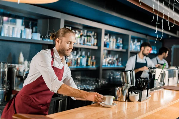 Handsome barista in apron serving coffee at wooden counter — Stock Photo