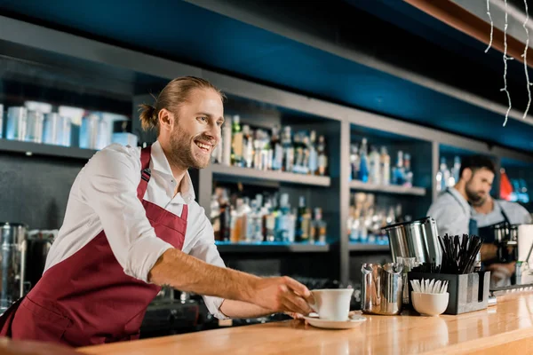 Beau barista souriant dans tablier servant du café au comptoir en bois — Photo de stock