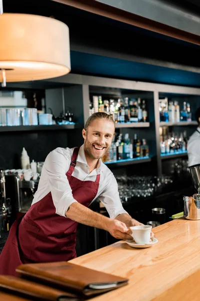 Adulto sonriente barista en delantal sirviendo café en mostrador de madera - foto de stock