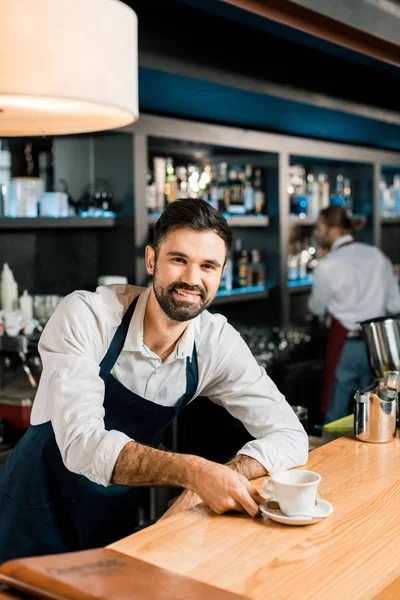 Cheerful smiling barista with coffee at wooden table — Stock Photo
