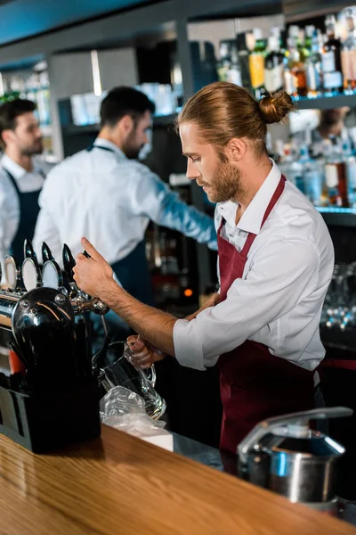 Barman em avental derramando cerveja em vidro atrás de balcão de madeira no bar — Fotografia de Stock