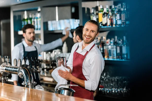 Barman sonriente pulido vidrio con tela en el mostrador de madera en el bar - foto de stock