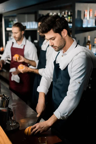 Handsome barman in apron slicing grapefruit with knife at counter — Stock Photo