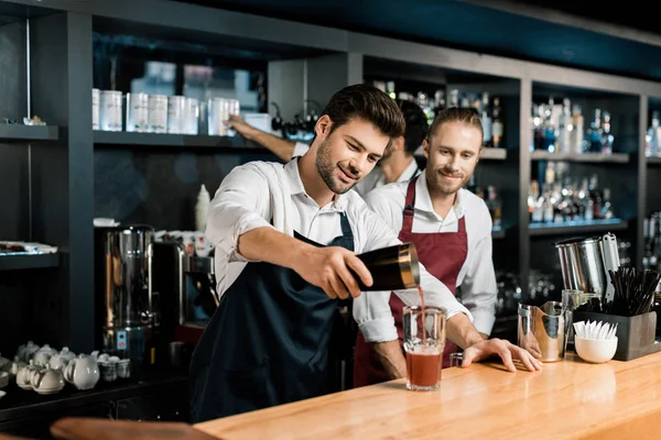 Дорослий бармен в фартусі наливає коктейль у склянку від шейкера за дерев'яною стійкою — стокове фото