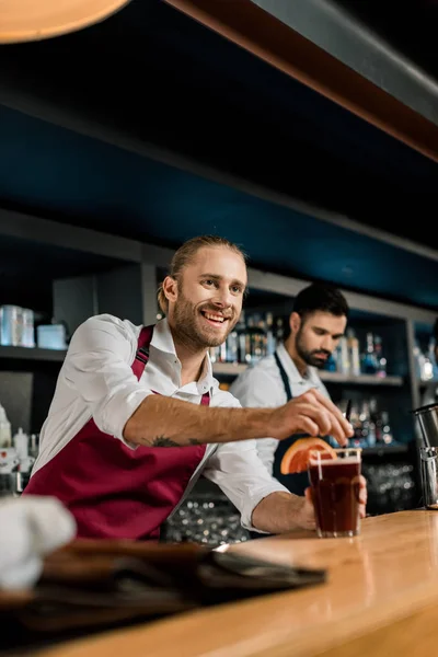 Schöner lächelnder Barkeeper, der Cocktails an der Holztheke serviert — Stockfoto