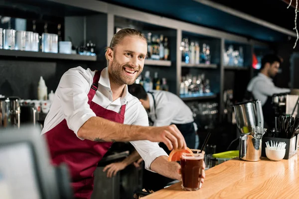 Smiling barman decorating cocktail with grapefruit slice at wooden counter — Stock Photo