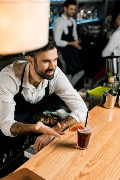 Guapo camarero sonriente que sirve cóctel con rodaja de pomelo y paja en el mostrador de madera - foto de stock