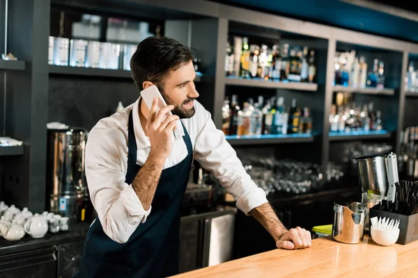 Barman talking on smartphone and standing in apron in bar — Stock Photo