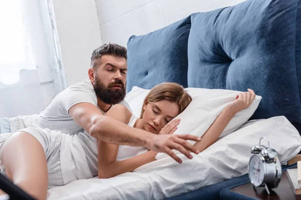 Sleepy bearded man waking up and turning off vintage alarm clock on nightstand while pretty girl sleeping in white pillows — Stock Photo