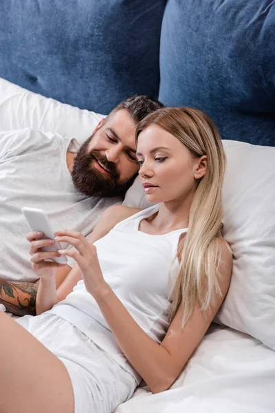 Barbu homme couché dans le lit et souriant tandis que jolie fille en utilisant smartphone le matin — Photo de stock