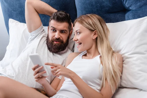 Joyeuse fille souriant et pointant du doigt sur l'écran du smartphone tandis que l'homme surpris barbu tenant la main sur la tête dans le lit — Photo de stock