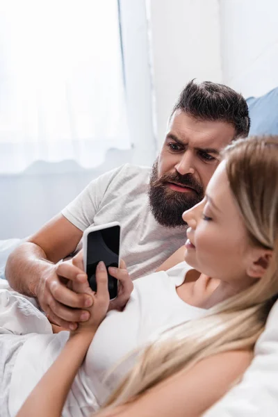 Pretty girl hiding smartphone screen from confused man in bed in morning — Stock Photo
