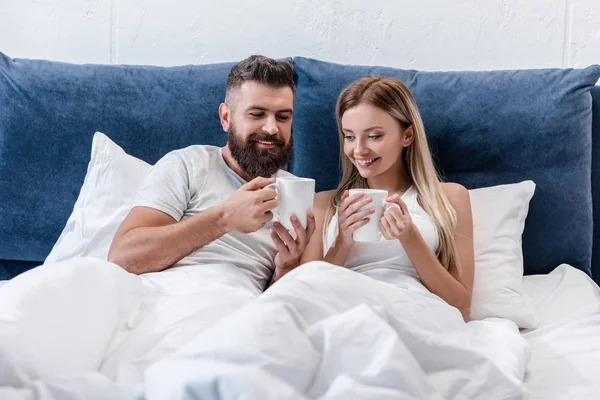 Verliebtes Paar liegt morgens im Bett und hält Kaffeetassen in der Hand — Stockfoto