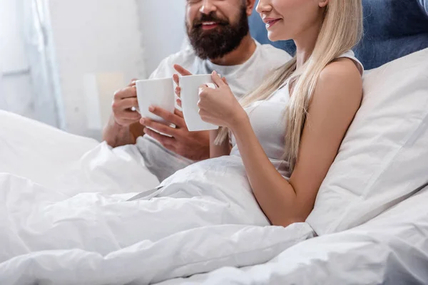 Vista cortada do casal apaixonado deitado na cama e segurando xícaras de café de manhã — Fotografia de Stock