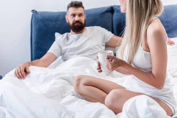 Бородатый мужчина серьезно смотрит на красивую молодую девушку с чашкой кофе в белой кровати — стоковое фото