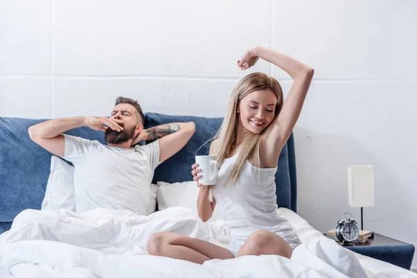 Бородатый мужчина зевает, в то время как привлекательная молодая женщина с чашкой кофе растягивается в постели — стоковое фото