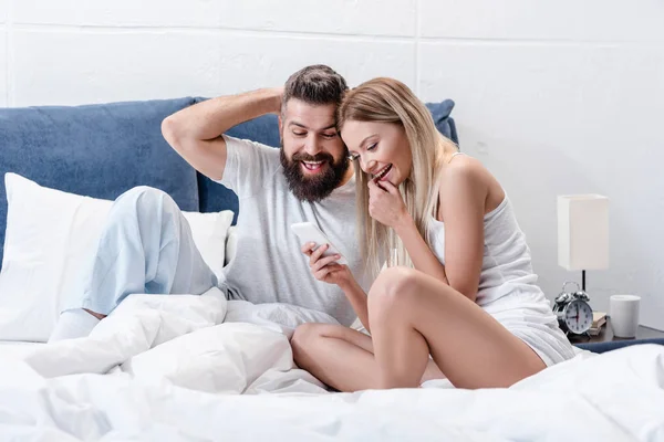 Happy young couple using smartphone and laughing in white bed in morning — Stock Photo
