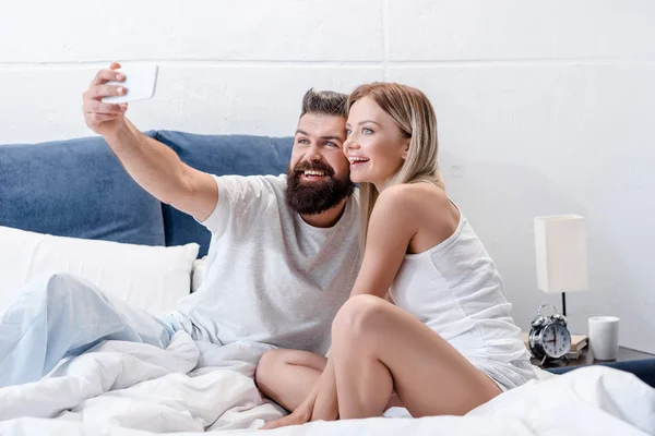 Bärtiger Mann und hübsches Mädchen lächeln und machen morgens ein Selfie im weißen Bett — Stockfoto