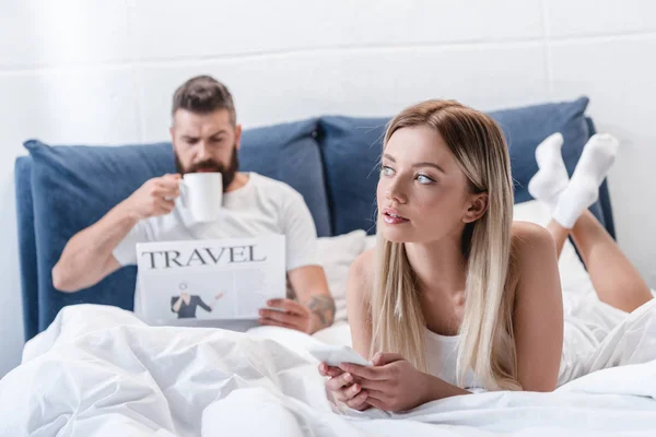 Chica bonita mintiendo y utilizando el teléfono inteligente mientras hombre barbudo beber café y leer el periódico de viajes en el dormitorio - foto de stock