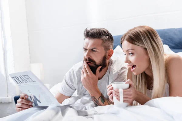 Sorprendido hombre y mujer joven con la taza acostada en la cama y leyendo el periódico de viajes - foto de stock