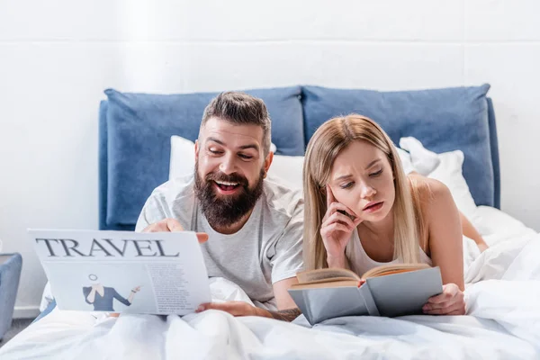 Fröhlicher Mann blickt auf Reisezeitung und nachdenkliche junge Frau liest Buch im Bett — Stockfoto