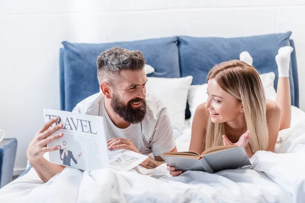 Joven barbudo con periódico de viaje y atractiva mujer con libro acostado en la cama y mirándose el uno al otro - foto de stock