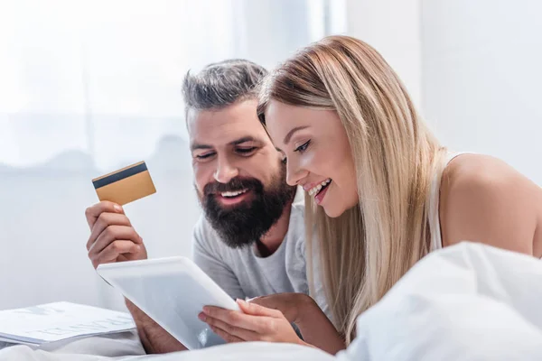 Casal atraente com tablet digital e cartão de crédito deitado no quarto brilhante — Fotografia de Stock