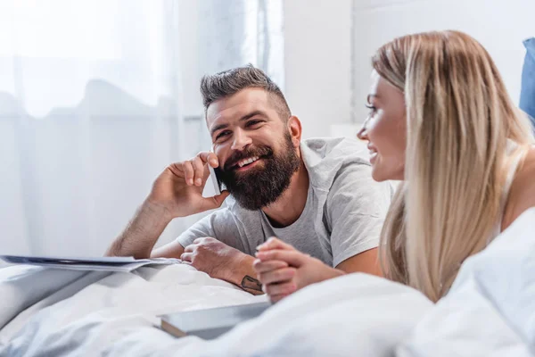 Barbu homme parler sur smartphone tandis que la fille blonde couché dans le lit — Photo de stock