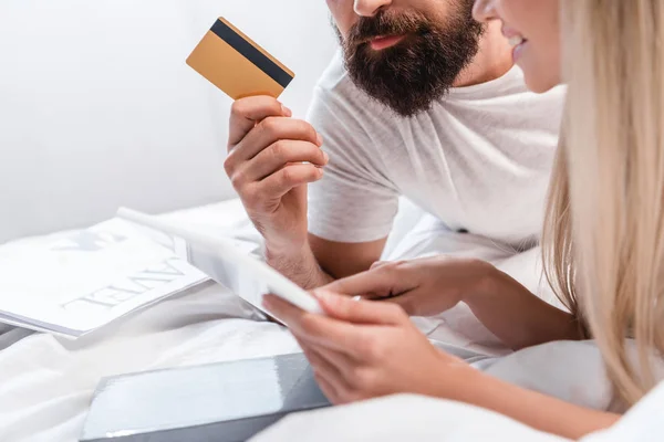 Young woman pointing with finger at digital tablet while bearded man holding credit card in bedroom — Stock Photo