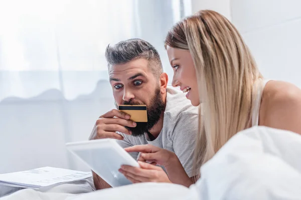 Joli couple avec carte de crédit et tablette numérique couché dans le lit et faire des achats en ligne — Photo de stock