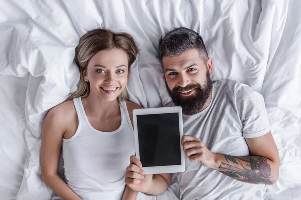 Couple souriant couché au lit, souriant et montrant la tablette numérique à l'appareil photo — Photo de stock