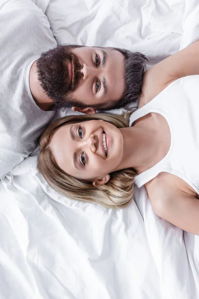 Hombre barbudo guapo y chica bonita tumbado cabeza a cabeza en la cama blanca y mirando a la cámara - foto de stock