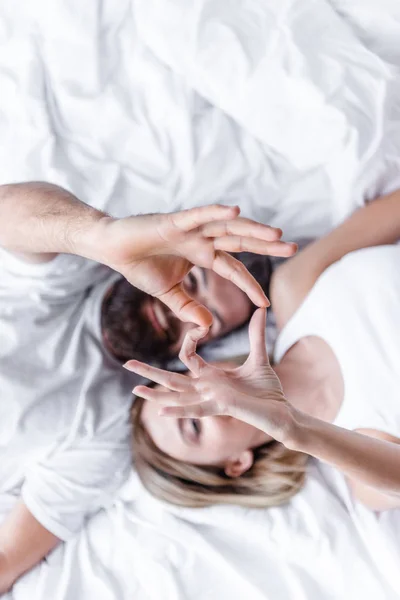 Pareja joven enamorada acostada cabeza a cabeza en la cama y mostrando el signo del corazón con los dedos - foto de stock