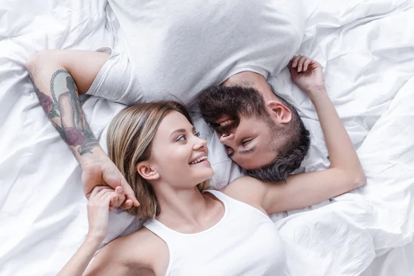 Vista superior del hombre guapo y la muchacha joven bonita que miente en la cama blanca, mirando el uno al otro y cogidas de la mano - foto de stock