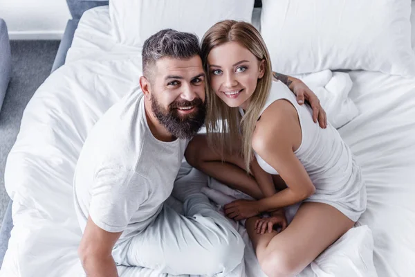 Blick von oben auf ein glückliches junges Paar, das im weißen Bett sitzt und in die Kamera schaut — Stockfoto