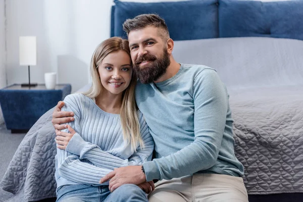 Glückliches junges Paar auf dem Boden sitzend, lächelnd und in die Kamera blickend im blauen Schlafzimmer — Stockfoto