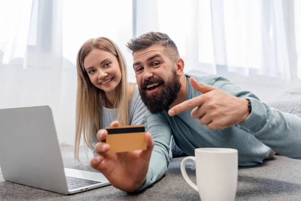 Cheerful couple lying on floor with laptop with credit card — Stock Photo
