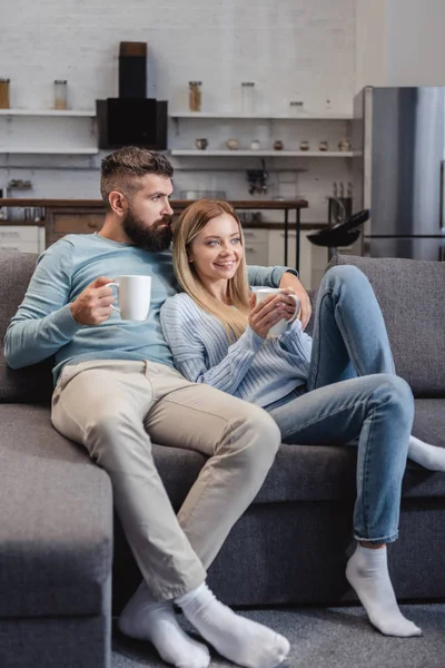 Cheerful couple sitting on sofa in casual clothing with cups — Stock Photo