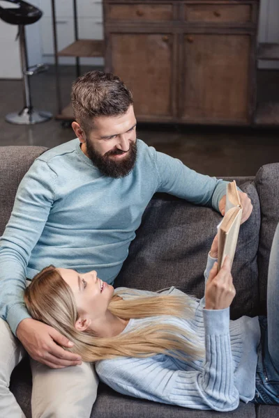 Cheerful wife holding book and lying on knees of husband — Stock Photo