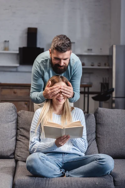 Cheerful husband closing eyes of happy wife with book — Stock Photo