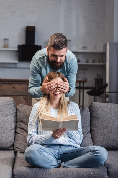 Alegre marido cerrando los ojos de feliz esposa con libro - foto de stock