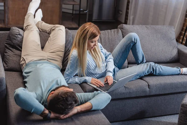 Husband lying on sofa and wife typing on laptop in room — Stock Photo