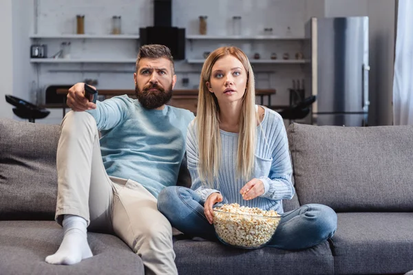 Sorprendida esposa sentada en el sofá con el marido y sosteniendo palomitas de maíz - foto de stock